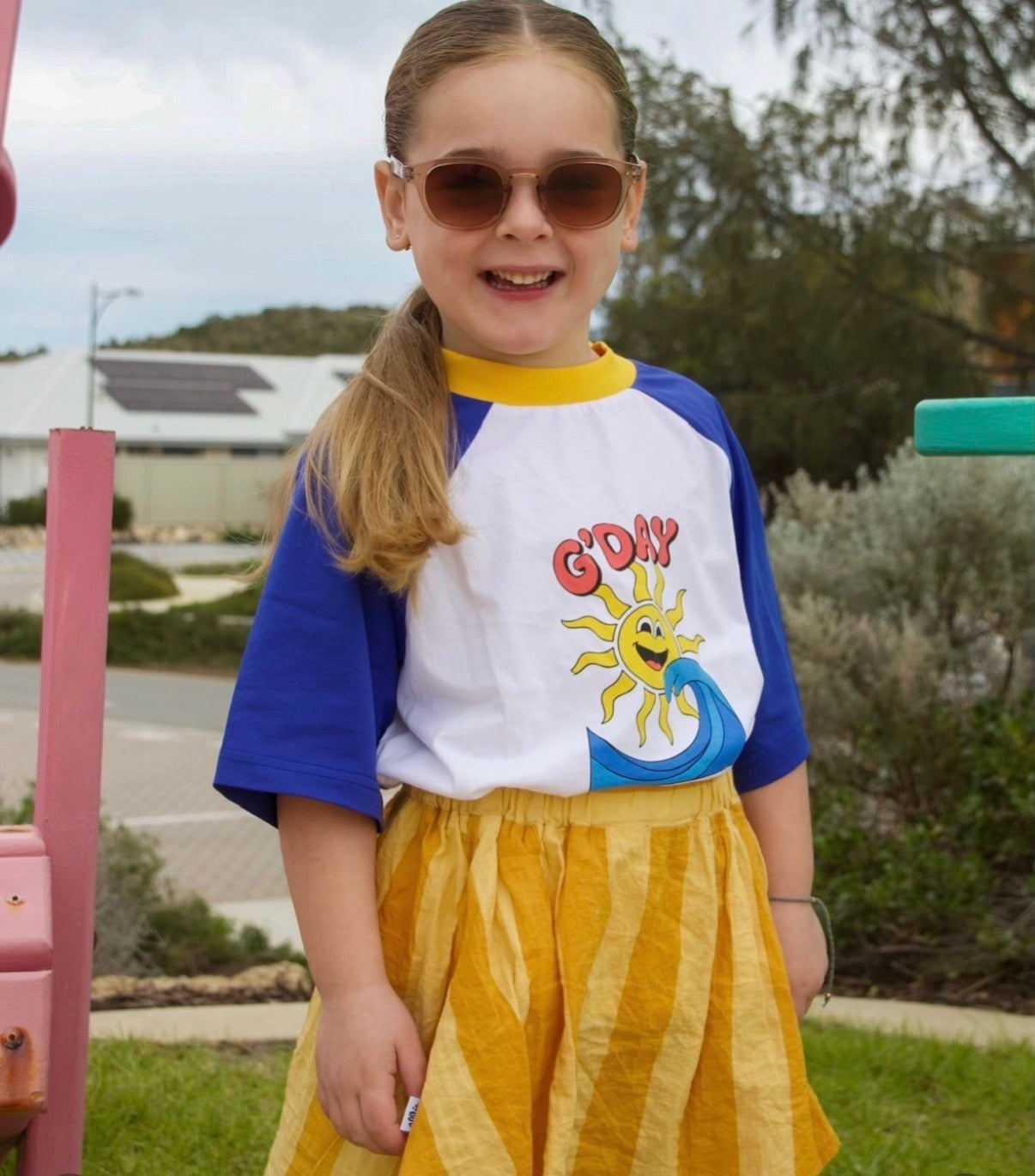 A young girl wearing a short-sleeve raglan t-shirt with blue and white panels, a yellow collar, and a 'G'DAY' sun graphic, paired with a yellow striped skirt, photographed outdoors in a park setting.