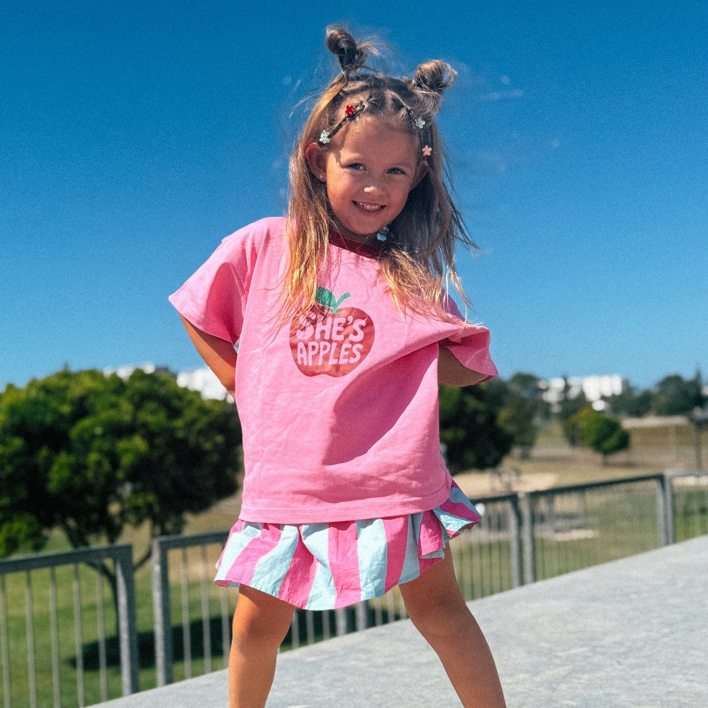A young girl with her hair in two buns stands barefoot on a concrete ledge outdoors under a clear blue sky. She is wearing a pink graphic T-shirt that says 'SHE'S APPLES' in a red apple outline, paired with a pink, white, and light blue striped skirt.
