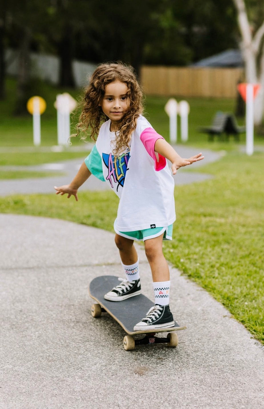 A young girl with curly brown hair stands on a skateboard on an outdoor paved path. She is wearing a white graphic t-shirt with pink and teal sleeves, teal shorts, white socks, and black sneakers.