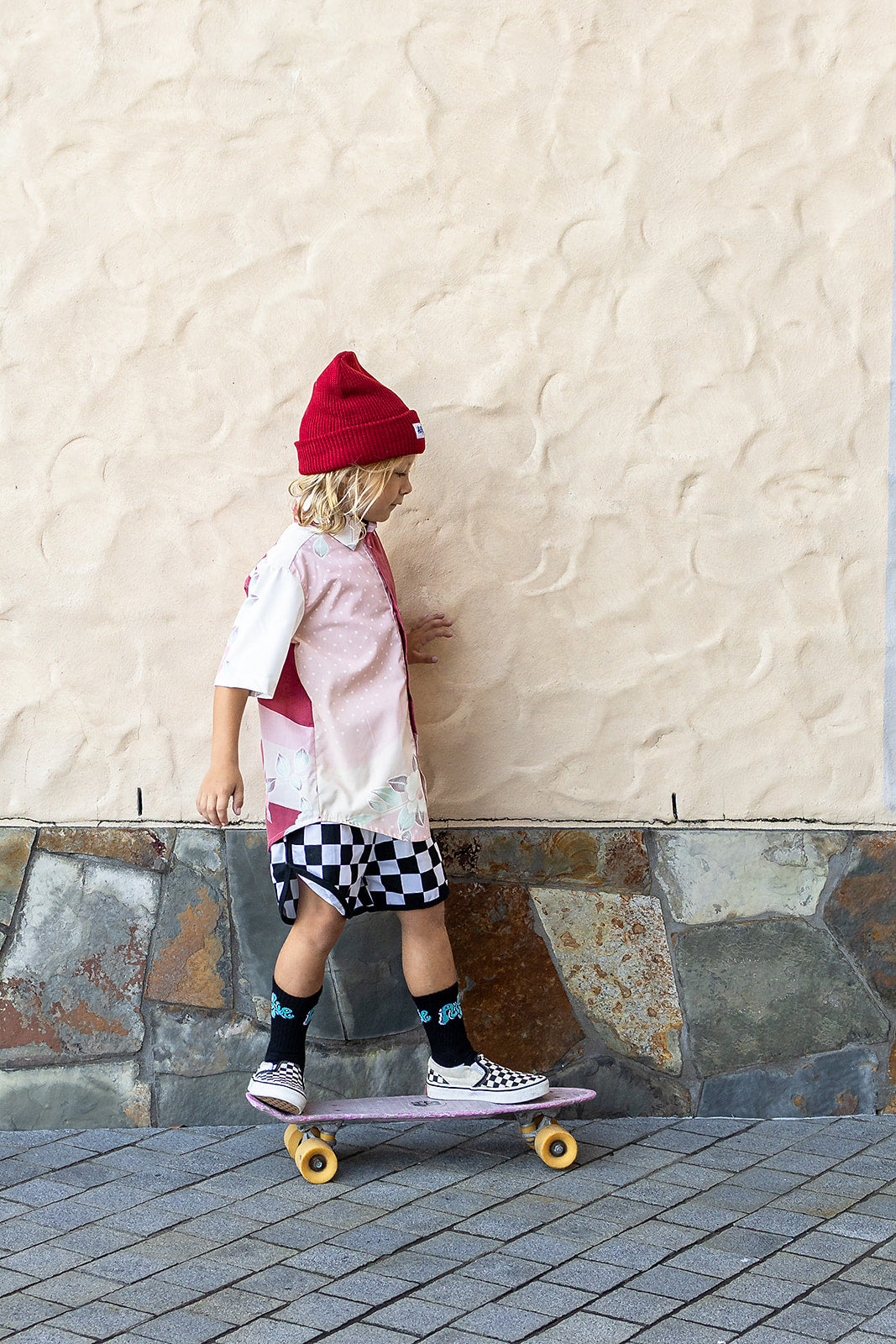 Child wearing a Alfie maroon beanie, Vintage Party Shirt and checkered shorts standing on a skateboard.