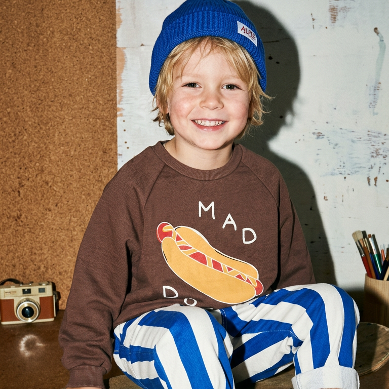 Child wearing a brown sweatshirt with a hot dog graphic and blue and white striped pants, sitting on a skateboard.