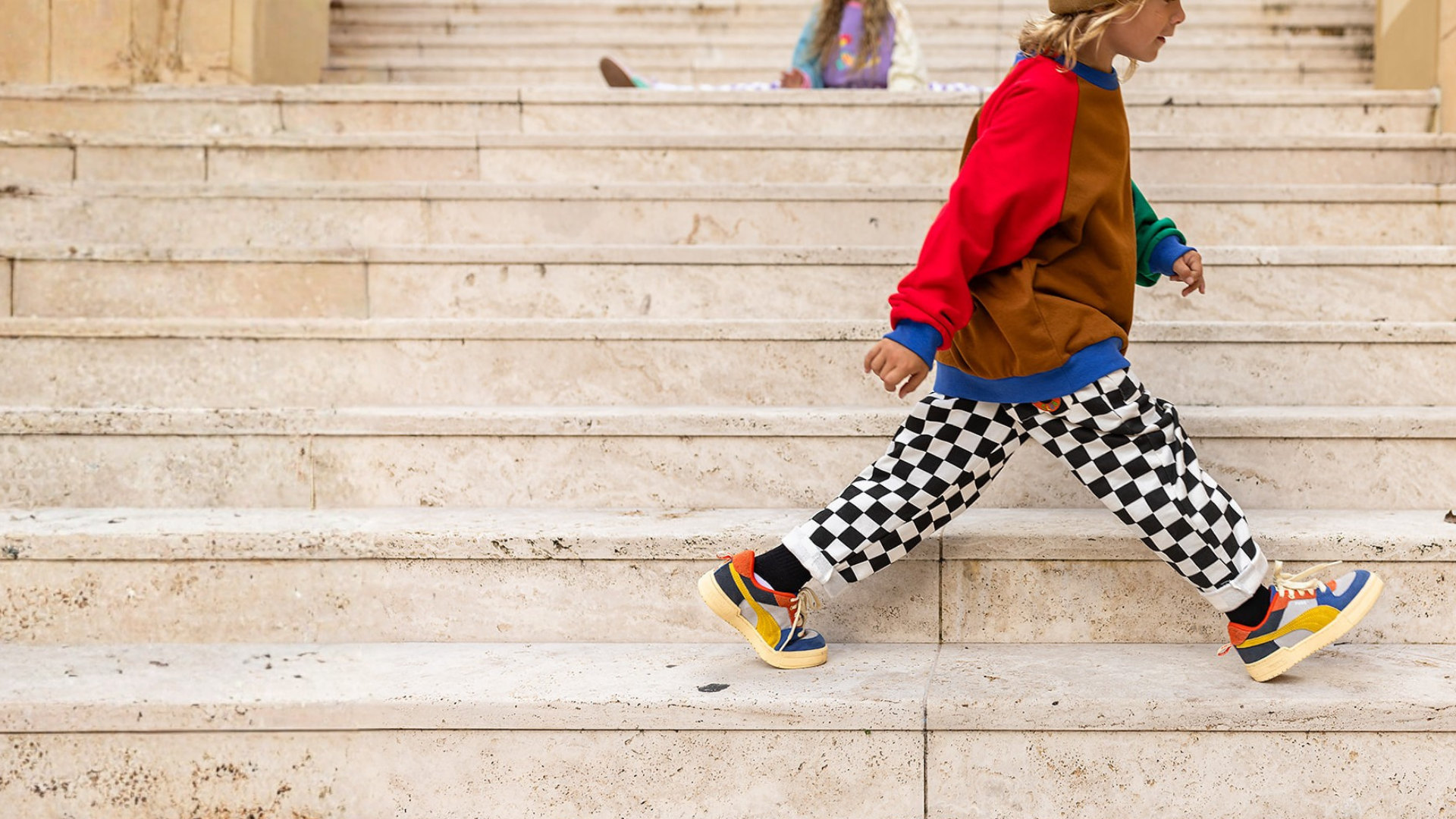 A child walking down wide stone steps, wearing a red and brown color-blocked sweatshirt, green and blue sleeves, black-and-white checkered pants, and colorful sneakers.